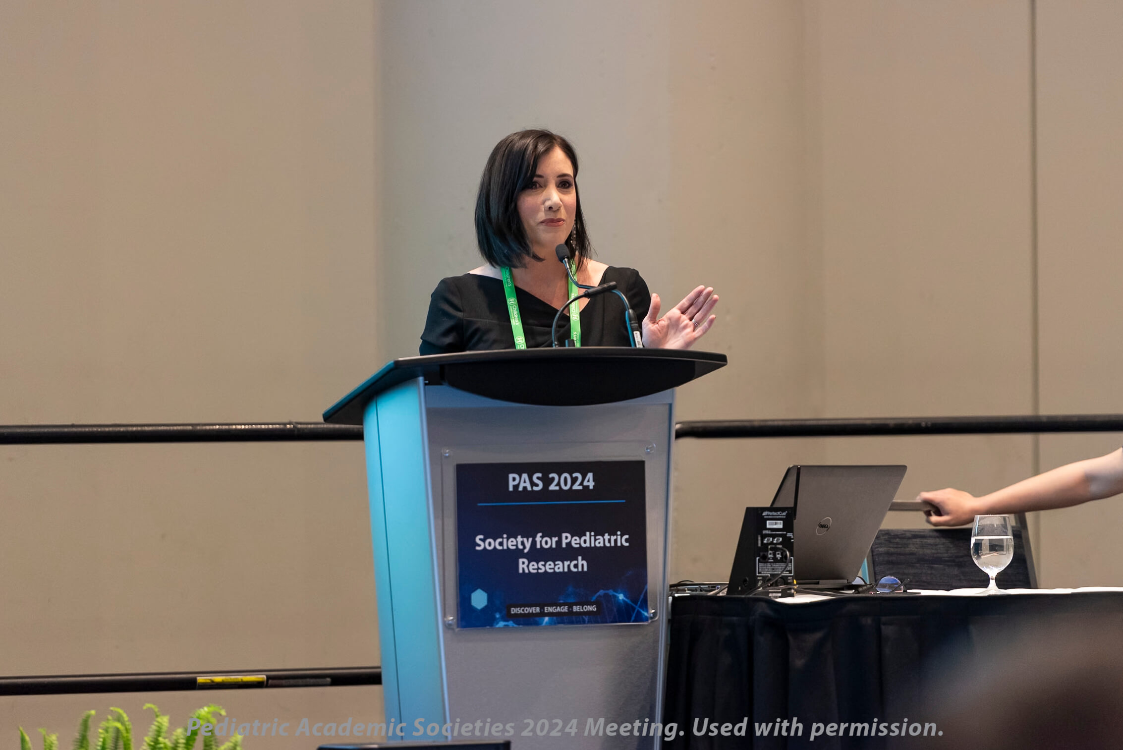 Woman in a black outfit with a green lanyard, speaking and gesturing at a podium labeled 'PAS 2024 Society for Pediatric Research,' with a table beside her holding a laptop, microphone, and glass of water, set against a plain wall and railing.
