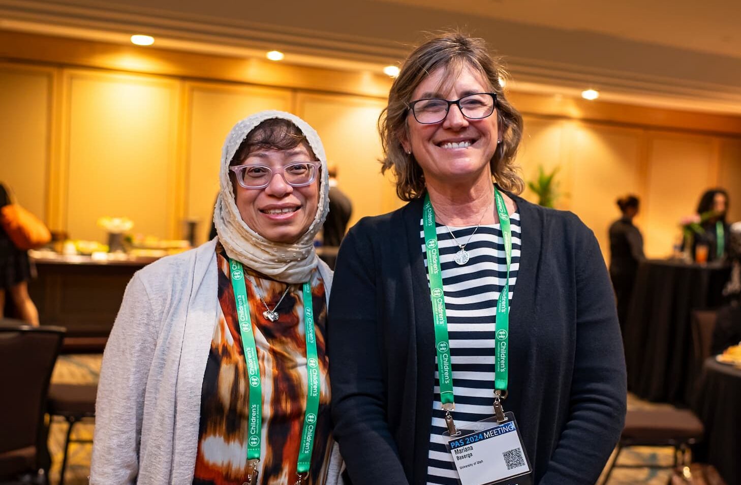 Two individuals standing together at a conference reception, wearing green lanyards and name badges, with tables and attendees visible in the background in a warmly lit room.