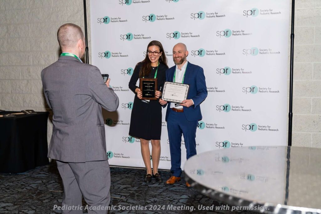 Two individuals standing in front of a backdrop with “Society for Pediatric Research” logos, each holding a recognition plaque and certificate, while another person takes a photo of them.