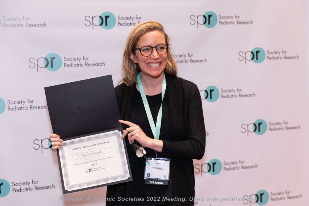 Person standing in front of a backdrop with “Society for Pediatric Research” logos, holding an open folder displaying a certificate.