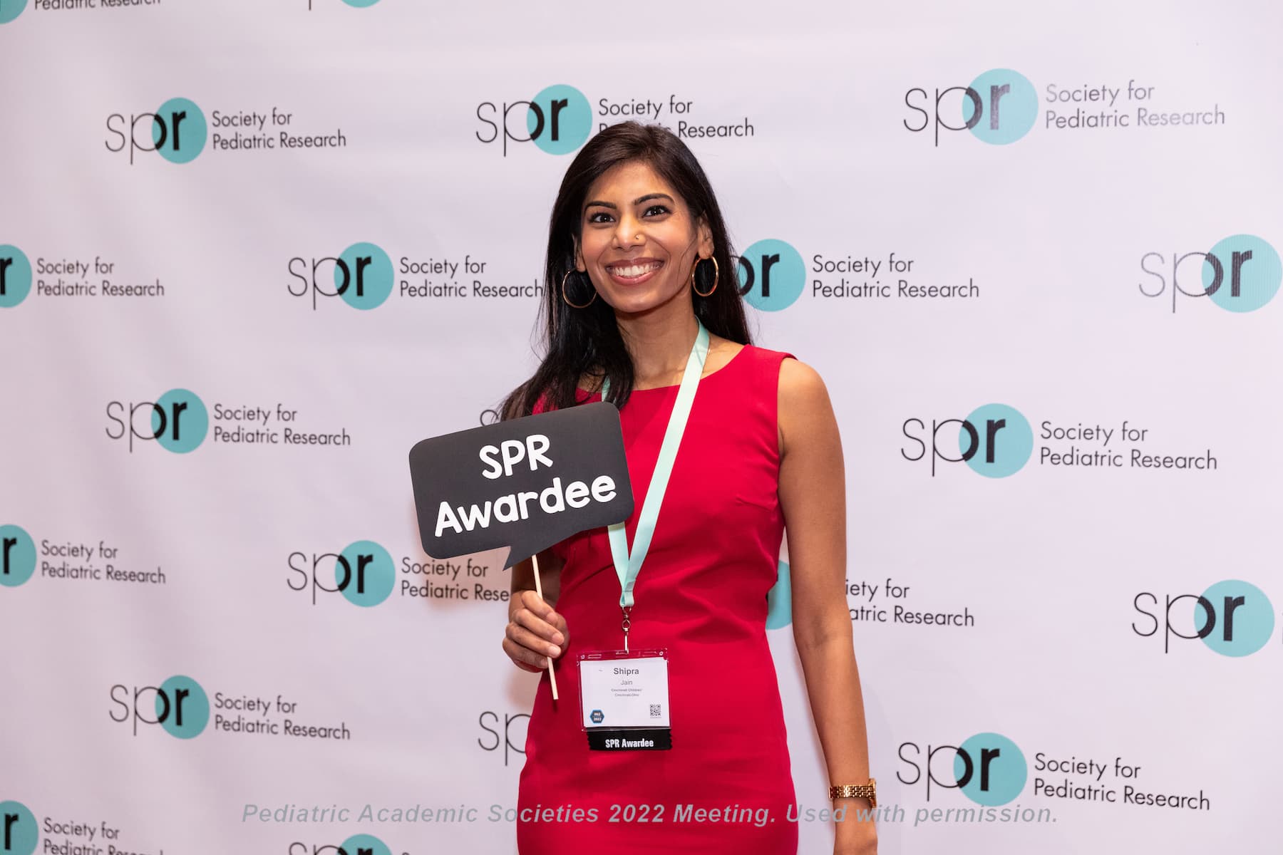 Person wearing a red dress and conference lanyard holding a sign that reads “SPR Awardee” in front of a backdrop with repeated Society for Pediatric Research logos at a Pediatric Academic Societies 2022 meeting.