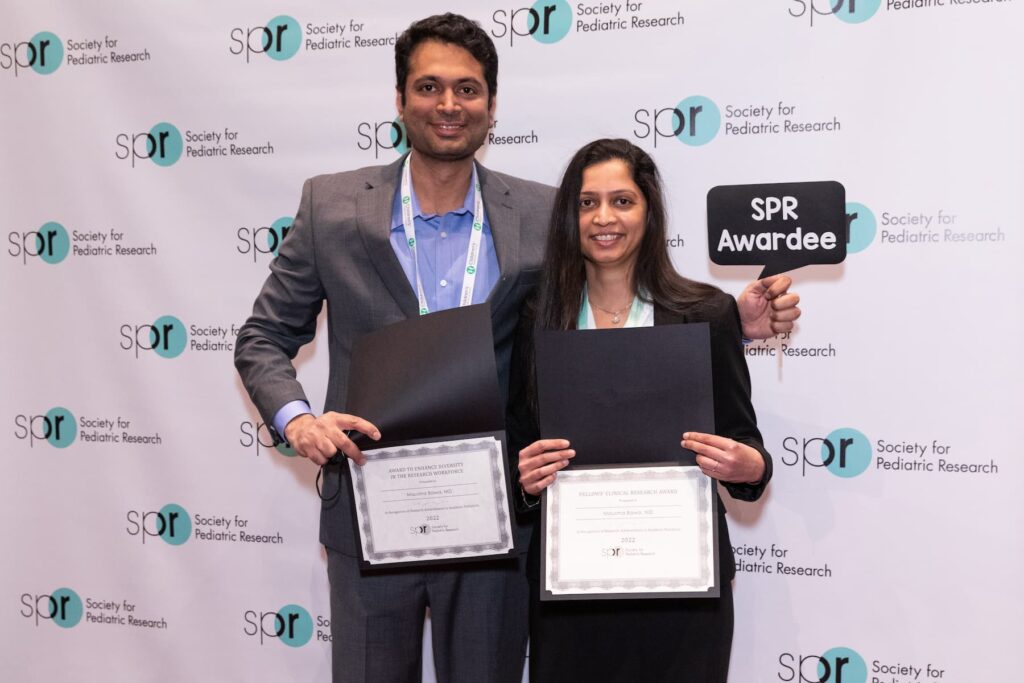 Two people standing in front of a backdrop with “Society for Pediatric Research” logos, holding certificates and a sign that reads “SPR Awardee.”