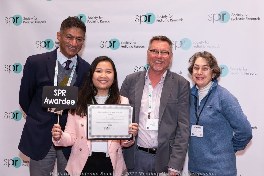 Group of five individuals standing in front of a backdrop with repeated Society for Pediatric Research logos; one person holds an award certificate and a sign reading “SPR Awardee,” while others wear conference badges and lanyards.
