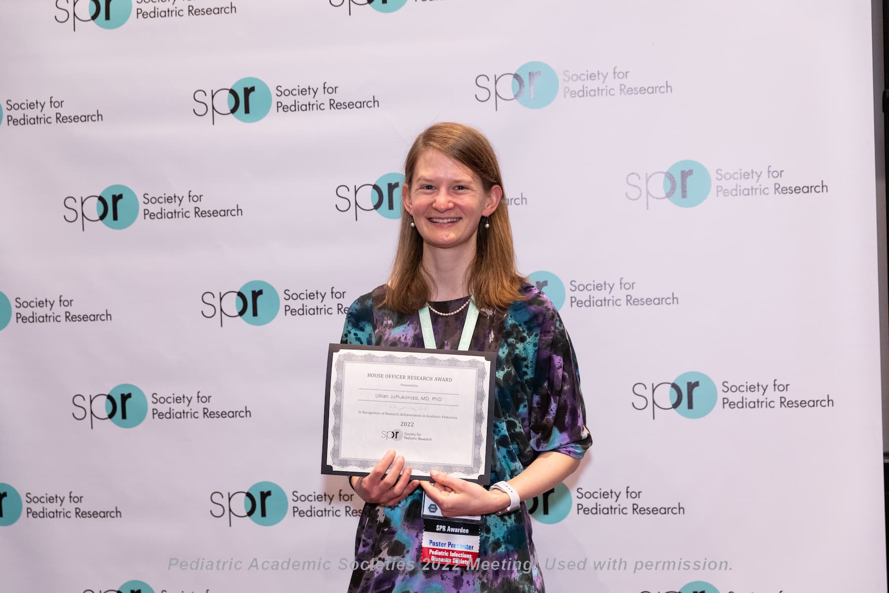 Person standing in front of a backdrop with repeated Society for Pediatric Research logos, holding an award certificate and wearing a conference lanyard at a Pediatric Academic Societies 2022 meeting.