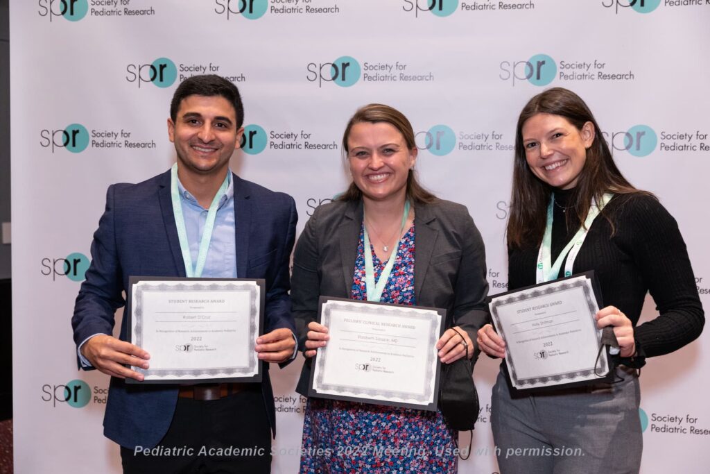 Three individuals standing in front of a backdrop with repeated Society for Pediatric Research logos, each holding an award certificate and wearing conference lanyards at a Pediatric Academic Societies 2022 meeting.