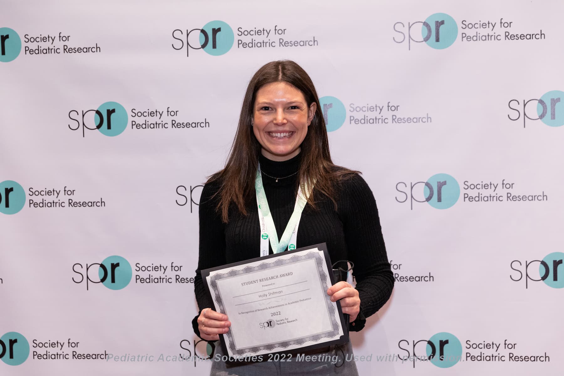 Person wearing a red dress and conference lanyard holding a sign that reads “SPR Awardee” in front of a backdrop with repeated Society for Pediatric Research logos at a Pediatric Academic Societies 2022 meeting.