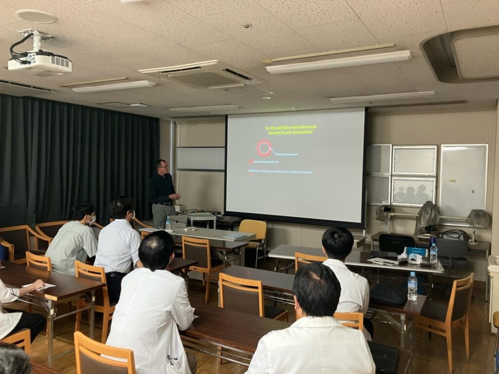 A classroom-style setting with several people seated at wooden tables facing a projector screen. The screen displays a presentation slide with a diagram and text in yellow and red. A presenter stands near the screen, and the room includes ceiling-mounted projectors, fluorescent lighting, and a mix of chairs and tables arranged for a lecture.