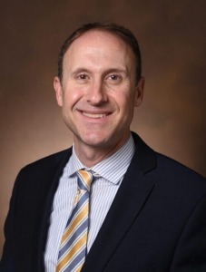 Person wearing a dark suit jacket, light blue dress shirt, and a striped yellow and gray tie, posed against a plain brown background.