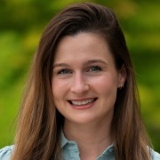 Person with long light brown hair wearing a light blue collared shirt, posed outdoors with a background of green foliage.