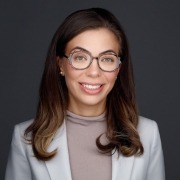 Person with long, wavy brown hair wearing a white blazer over a light high-neck top, posed against a dark gray background.