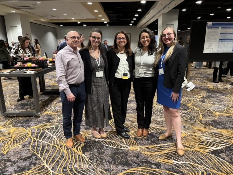 Five people standing together in a conference setting with patterned carpet, wearing business attire and name badges. Behind them are tables with refreshments and a poster board displaying research content, with other attendees visible in the background.