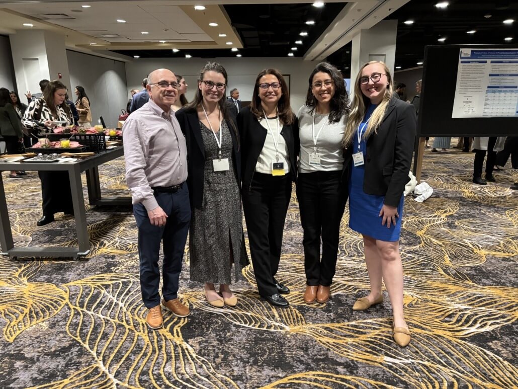 Five people standing together in a conference setting with patterned carpet, wearing business attire and name badges. Behind them are tables with refreshments and a poster board displaying research content, with other attendees visible in the background.