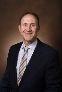 Person wearing a dark suit jacket, light blue dress shirt with thin vertical stripes, and a striped tie in shades of yellow, blue, and white, smiling at the camera against a brown gradient background.