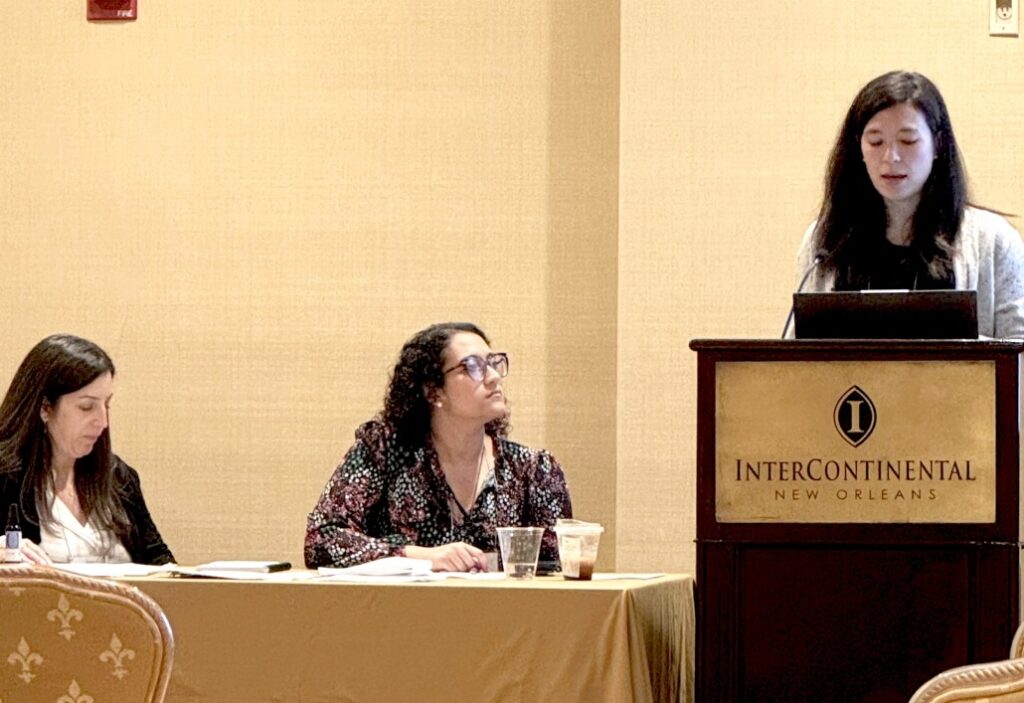 Two individuals standing indoors during an award presentation. One person is holding a wooden plaque with a gold emblem and engraved text that reads: “Presented to Amy Hair, M.D. In appreciation for your service as President of the Southern Society for Pediatric Research 2024–2025.” Both are wearing business attire and conference name badges, and a microphone is visible in the foreground.