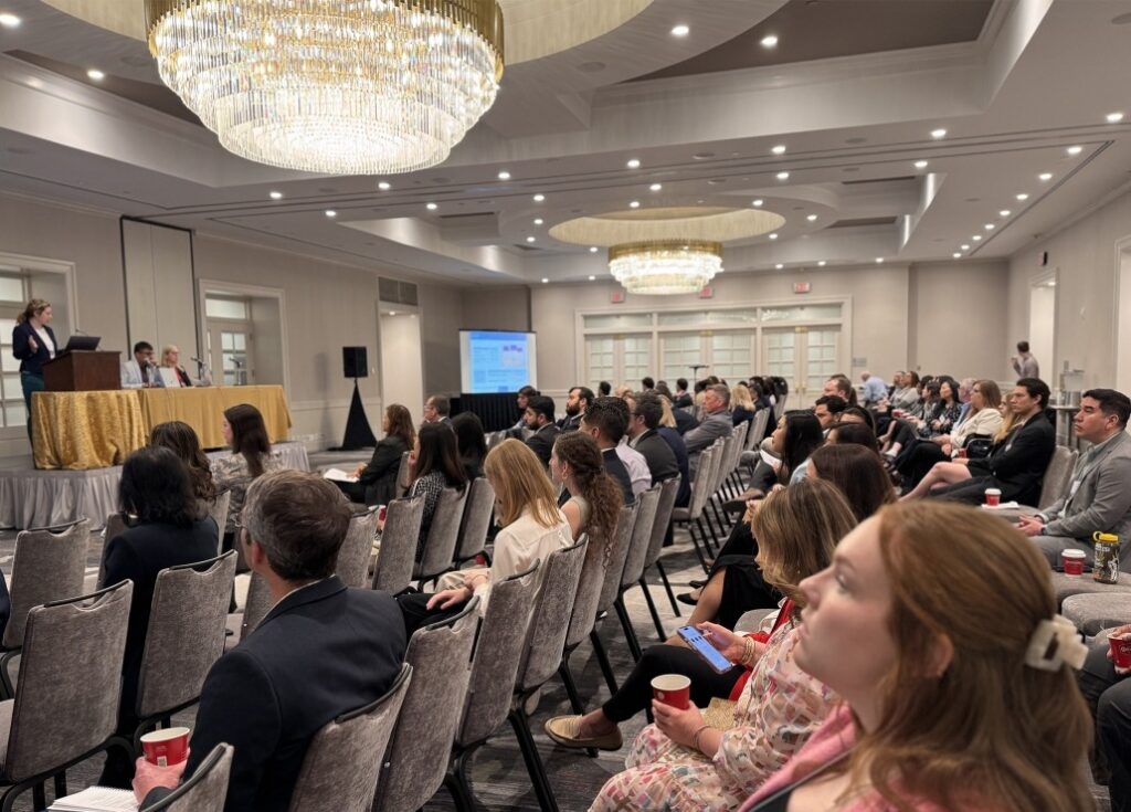 A conference room filled with attendees seated in rows facing a stage. On the stage, a speaker stands at a podium with three panelists seated beside a table covered in a gold cloth. Two large chandeliers hang from the ceiling, and a projection screen displays content in the background. The setting is formal, with participants dressed in business attire.