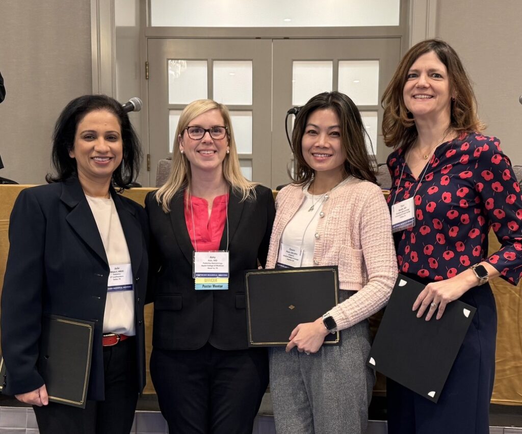Four individuals standing indoors in front of a set of double doors, each wearing business attire and conference name badges. Two of them are holding black award folders, suggesting a recognition or award ceremony setting. A microphone is visible on the left side of the image.