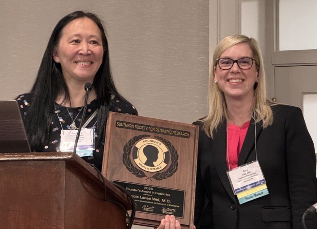 Two individuals standing indoors, one holding a wooden plaque that reads “Southern Society for Pediatric Research – Abbott Nutrition Pediatric Educator Award” with the recipient’s name engraved. The plaque features a circular emblem with laurel design. Both are wearing business attire and conference name badges, and a microphone is visible in the foreground.