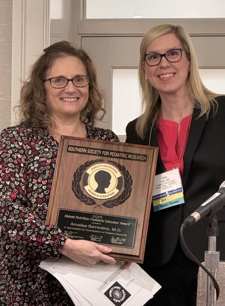 Two individuals standing indoors, one holding a wooden plaque that reads “Southern Society for Pediatric Research – Abbott Nutrition Pediatric Educator Award” with the recipient’s name engraved. The plaque features a circular emblem with laurel design. Both are wearing business attire and conference name badges, and a microphone is visible in the foreground.
