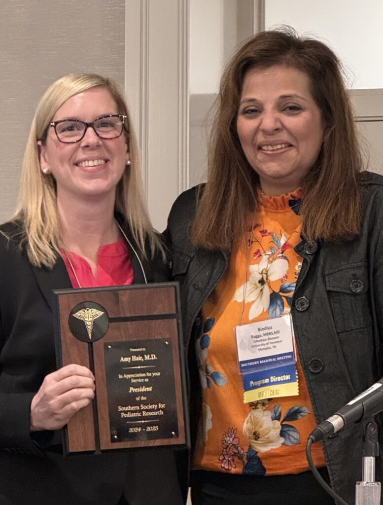 Two individuals standing indoors during an award presentation. One person is holding a wooden plaque with a gold emblem and engraved text that reads: “Presented to Amy Hair, M.D. In appreciation for your service as President of the Southern Society for Pediatric Research 2024–2025.” Both are wearing business attire and conference name badges, and a microphone is visible in the foreground.