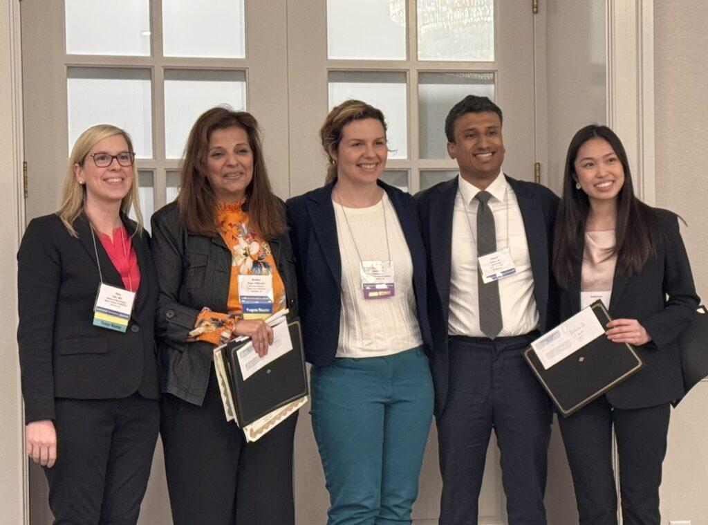 Five individuals standing indoors in front of a set of glass-paneled double doors, dressed in business attire and wearing conference name badges. Two people are holding black award folders, and one person is holding multiple documents, suggesting an award or recognition event.