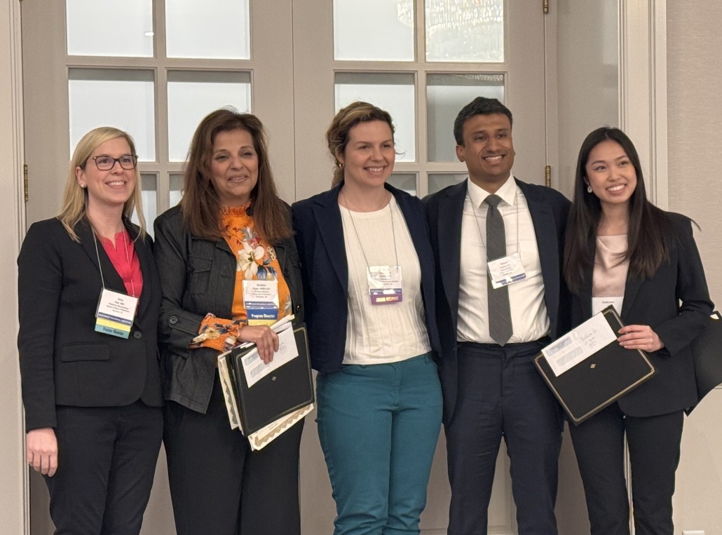 Five individuals standing indoors in front of a set of glass-paneled double doors, dressed in business attire and wearing conference name badges. Two people are holding black award folders, and one person is holding multiple documents, suggesting an award or recognition event.