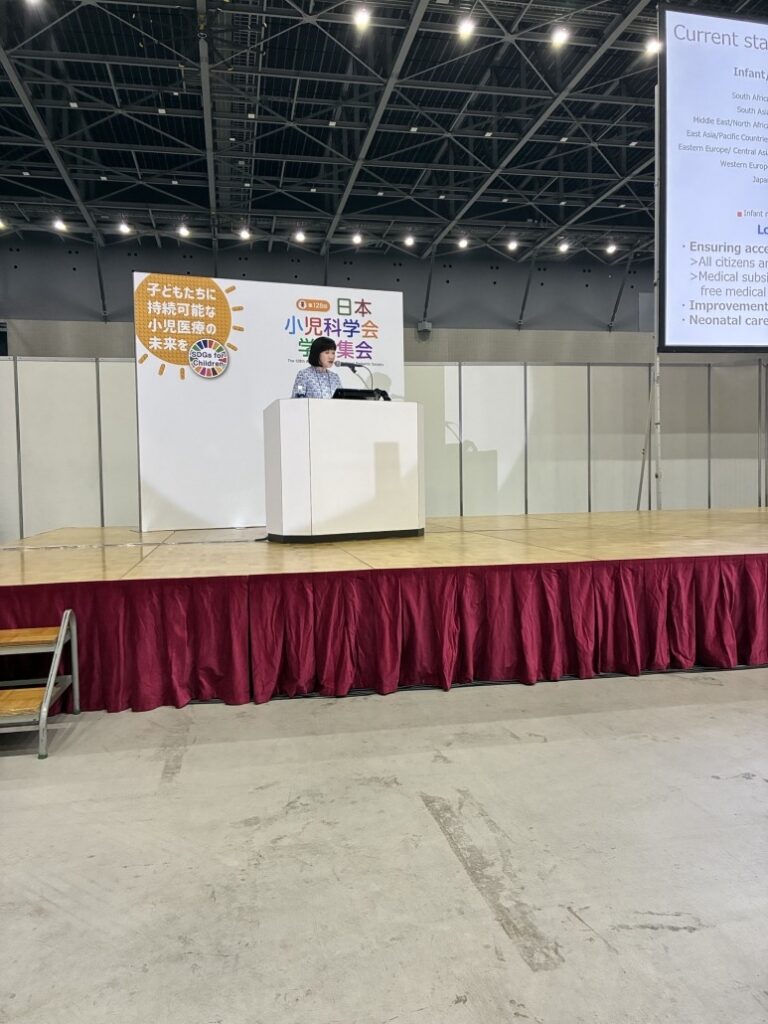 Speaker standing at a podium on a stage with a red skirted front, delivering a presentation at a conference. Behind the podium is a backdrop with Japanese text and colorful signage, and a large screen to the right displays a slide titled “Current status” with bullet points about infant health and neonatal care. The venue has high ceilings with visible lighting fixtures.