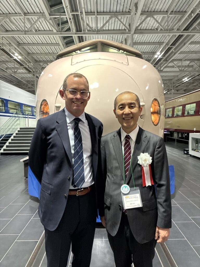 Two individuals in formal suits standing in front of a large vintage train engine inside a modern exhibition hall with high ceilings and multiple train cars displayed in the background.