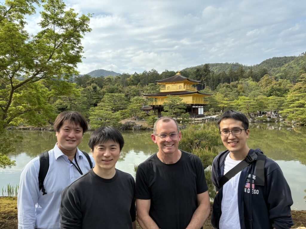 Four people standing outdoors in front of a scenic pond with lush greenery and the Golden Pavilion (Kinkaku-ji) temple in the background. The temple is a gold-colored structure surrounded by trees and reflected in the water, with mountains visible in the distance under a partly cloudy sky.