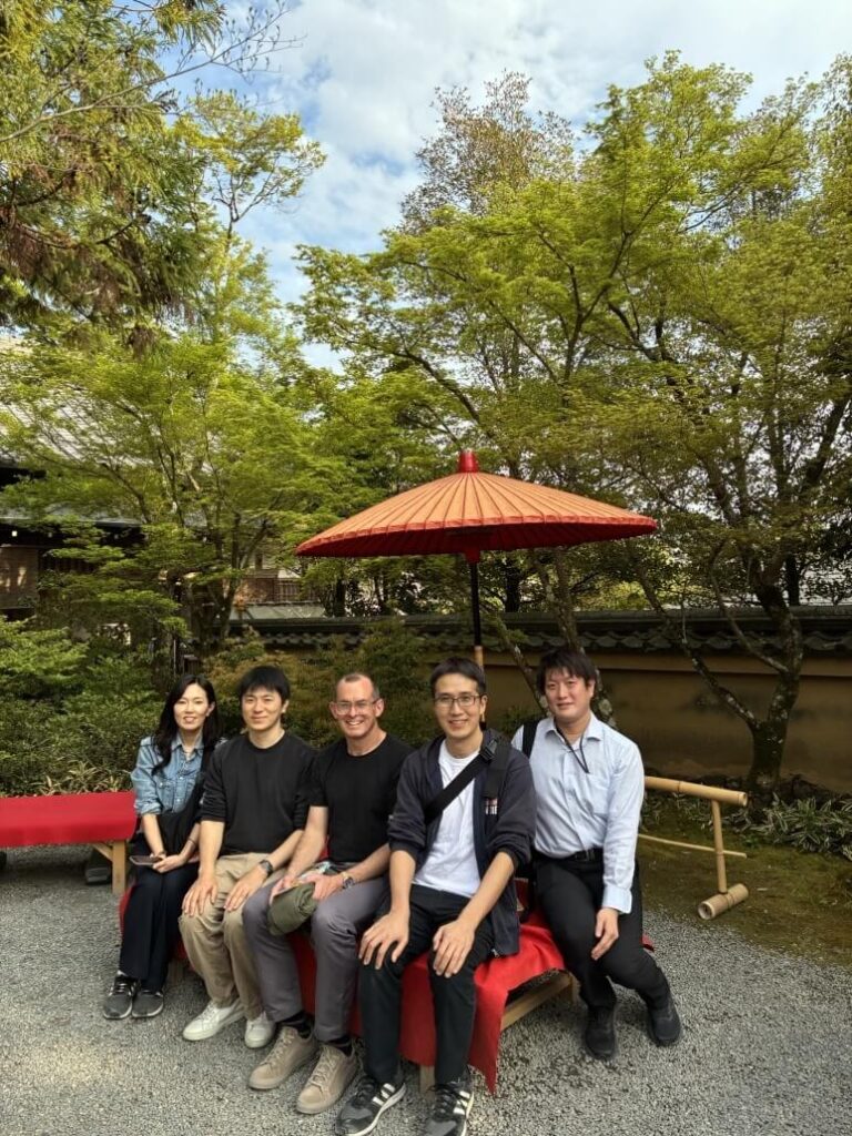Five people sitting on a red bench outdoors under a traditional Japanese umbrella in a garden setting. The background features lush green trees, a stone pathway, and a wooden fence, creating a serene and natural atmosphere.