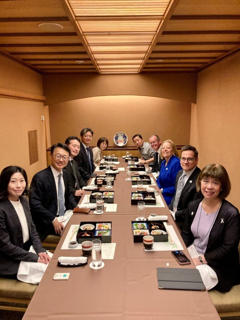A formal dining setting in a private room with a long rectangular table, where multiple people are seated on both sides. The table is set with bento-style meals, cups, glasses of water, and menus, creating an organized and elegant atmosphere.