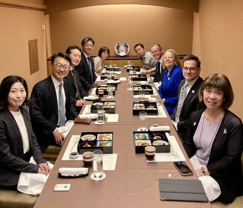 A group of people seated around a long table in a traditional-style dining room, with neatly arranged bento boxes, cups, and glasses of water in front of them. The table is covered with a brown cloth, and the room features warm beige walls and a decorative plate on the back wall.