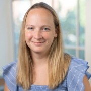 Person with straight, shoulder-length blonde hair wearing a light blue top with ruffled sleeves, posed indoors with a softly blurred background featuring windows and greenery.