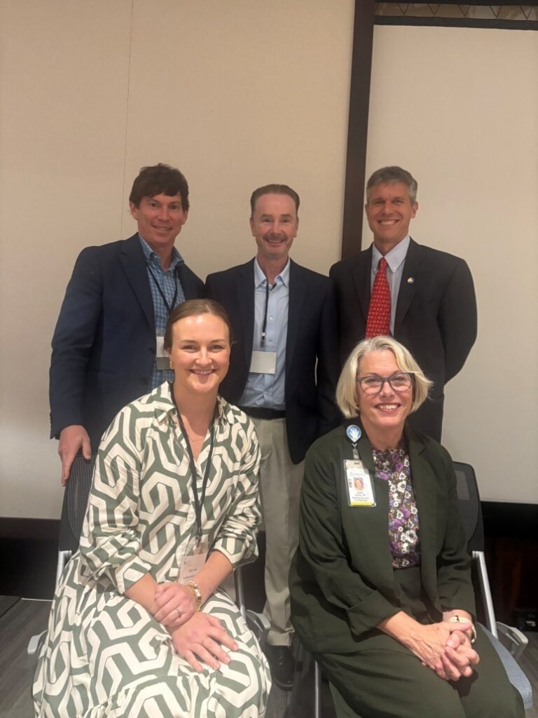 Group photo of five individuals in professional attire at an indoor event. Three people are standing in the back wearing suits and name badges, while two are seated in front, one in a patterned dress and the other in a dark outfit with a visible ID badge. Background shows a beige wall and part of a projection screen