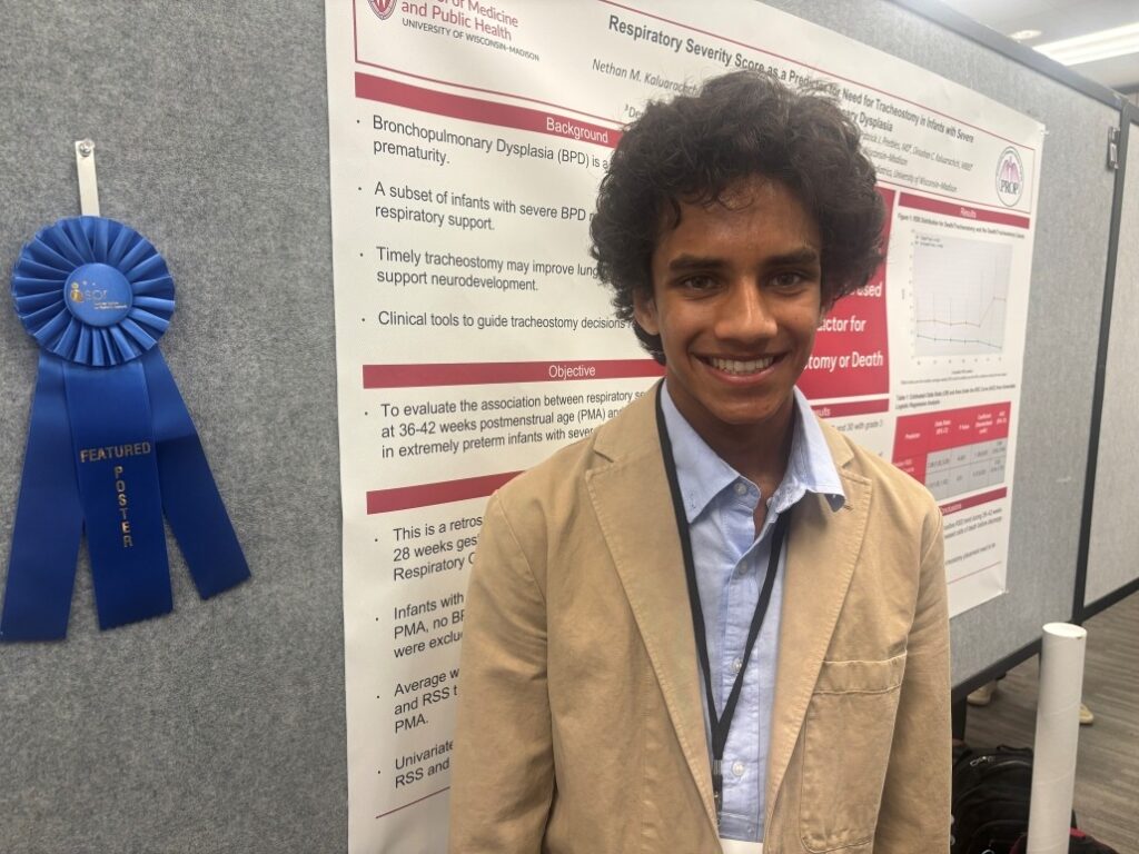 Person standing in front of a scientific research poster titled “Respiratory Severity Scores & Predictors for Tracheostomy,” featuring text, charts, and a blue ribbon labeled “Featured Poster” attached to the display board.
