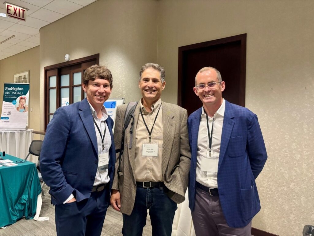 Three individuals standing together at an indoor conference reception area. They are wearing business attire with name badges and lanyards. Behind them is a beige wall with two dark wooden doors and a sign for 'ProNephro aNT (NGAL)' on a table with a teal tablecloth. An exit sign is visible above the doorway