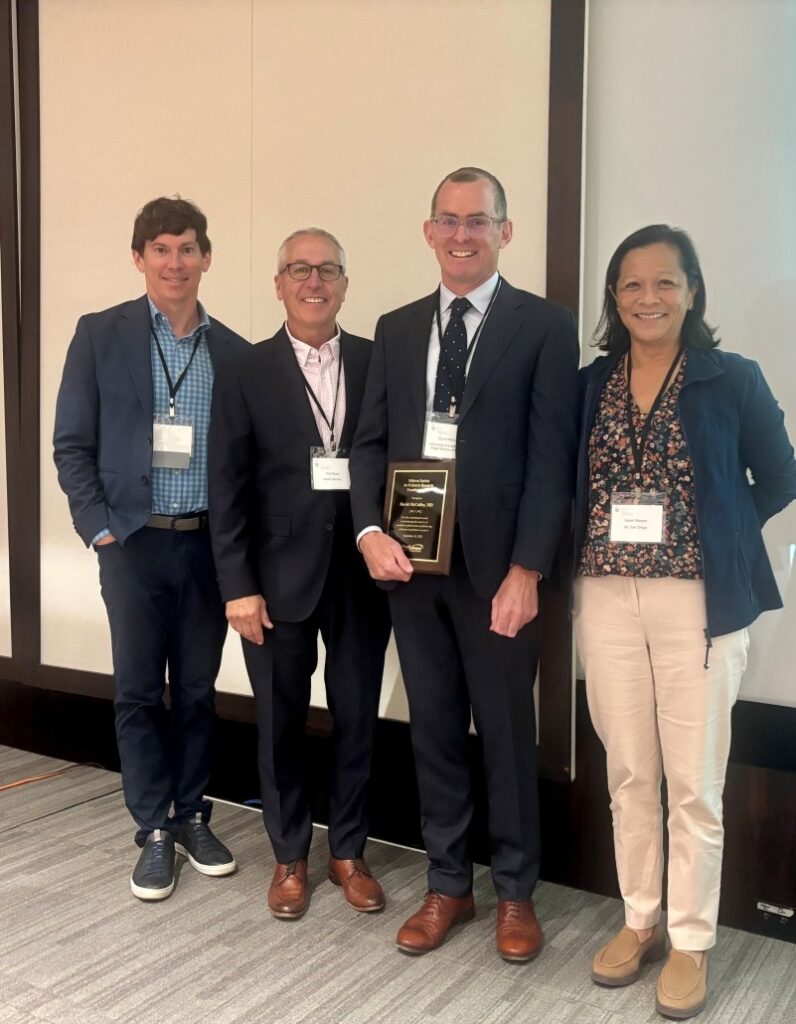 Four individuals standing indoors at a conference event. All are wearing business attire with name badges. One person in the center is holding a rectangular award plaque. Background includes beige walls and a projection screen