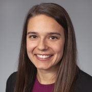 Person with straight, long brown hair wearing a black blazer over a dark magenta top, posed against a plain gray background.