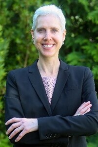 Person with short white hair wearing a black blazer over a light lace top, standing outdoors with arms crossed in front of green foliage.