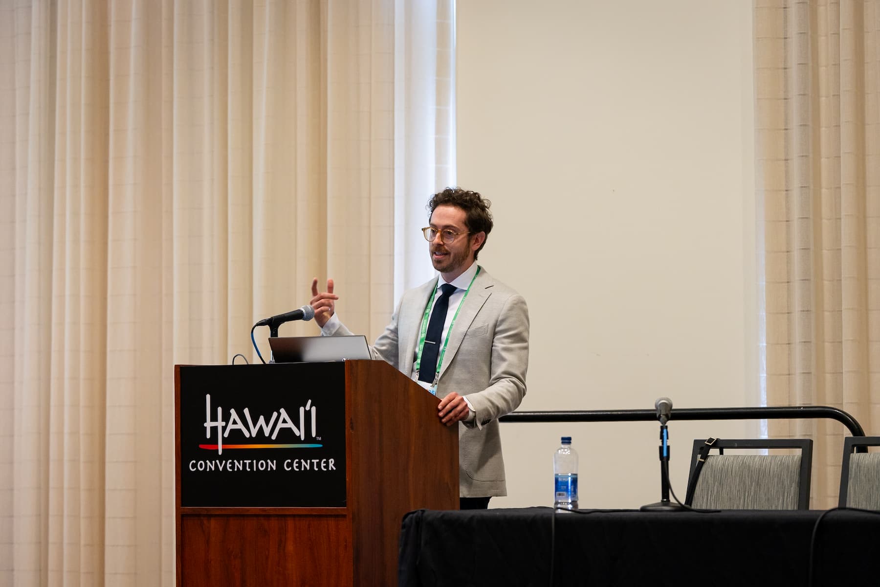 Speaker presenting at a podium labeled “Hawai‘i Convention Center” during a conference, wearing a light-colored suit and green lanyard, with a microphone, laptop, and water bottle visible on the table beside the podium.