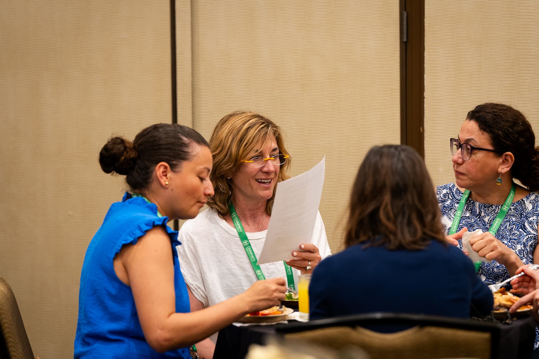 Four individuals seated around a table at a conference, wearing green lanyards and name badges, with one person holding a sheet of paper while others have plates of food and drinks in front of them.