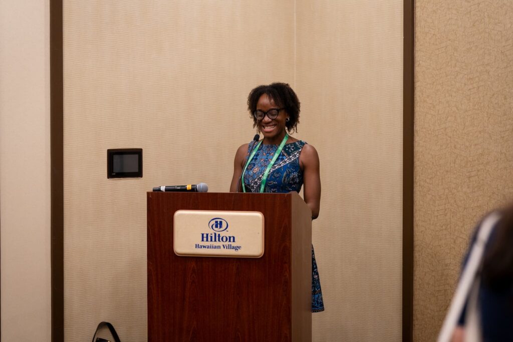 Person standing at a podium labeled “Hilton Hawaiian Village” with a microphone and conference badge visible.
