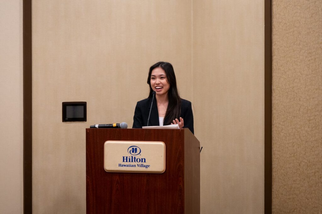 A person standing behind a wooden podium with a Hilton Hawaiian Village logo, delivering a presentation in a conference room. A microphone and papers are placed on the podium, and the background features beige walls.