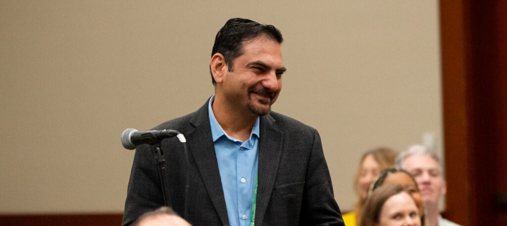 A person standing near a microphone in a conference room, holding papers and a drink. Several other attendees are seated in rows of chairs, wearing event lanyards. The setting appears to be a formal discussion or Q&A session.