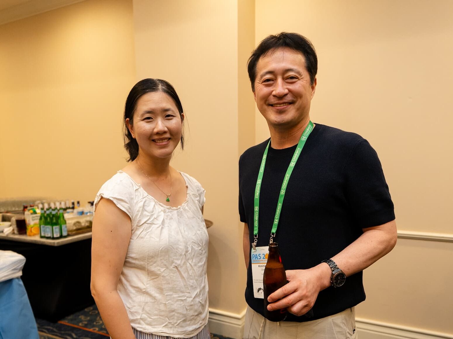 Two individuals standing together at a conference social event, wearing green lanyards and name badges; one person is holding a beverage bottle, with a table of drinks and snacks visible in the background.