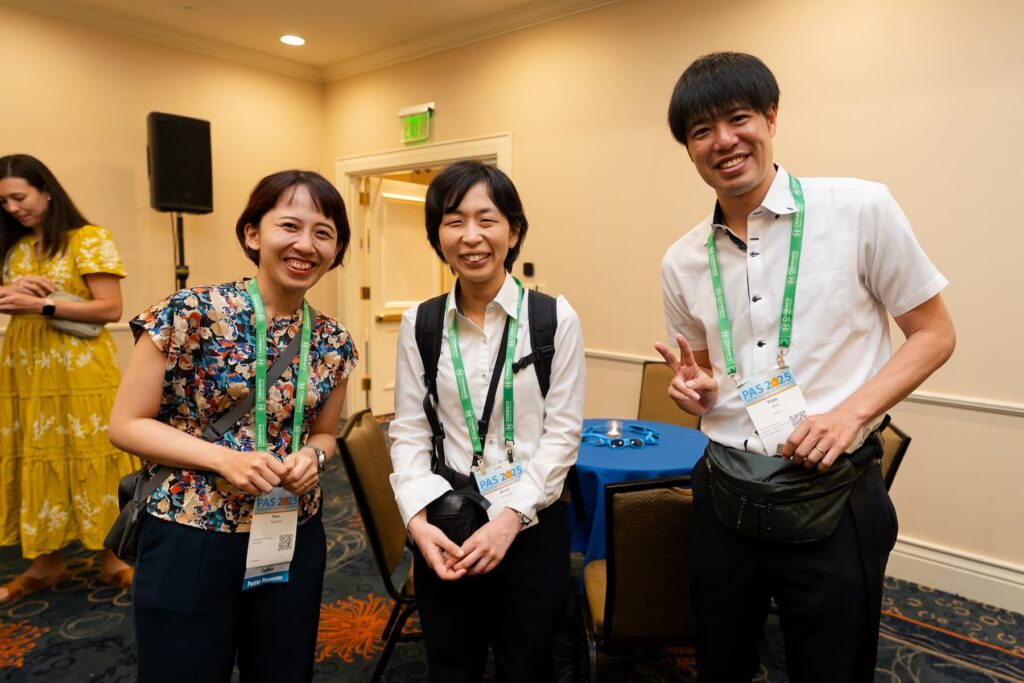 Three individuals standing together at a conference networking event, wearing green lanyards and name badges; one person is making a peace sign gesture. A round table with a blue tablecloth and chairs is visible behind them, along with beige walls and a door in the background.