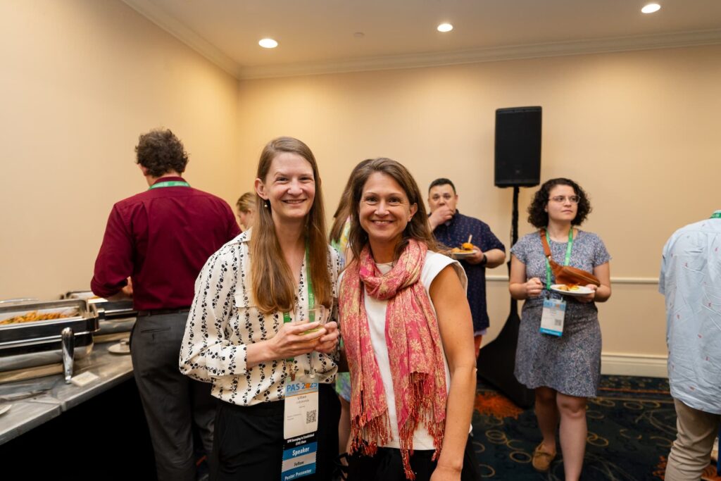 Two individuals standing together at a buffet-style event, holding food and drinks. Behind them, a table with chafing dishes containing food is visible, and several other attendees are serving themselves or eating. The setting appears to be an indoor conference or networking reception with cream-colored walls and dark carpet.