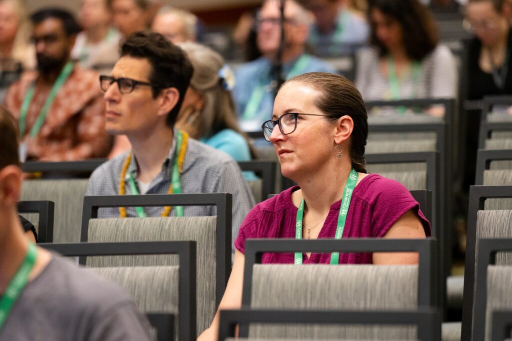 Audience members seated in rows of chairs at a conference or seminar, wearing event lanyards. The setting appears to be an indoor meeting room with multiple attendees visible in the background.