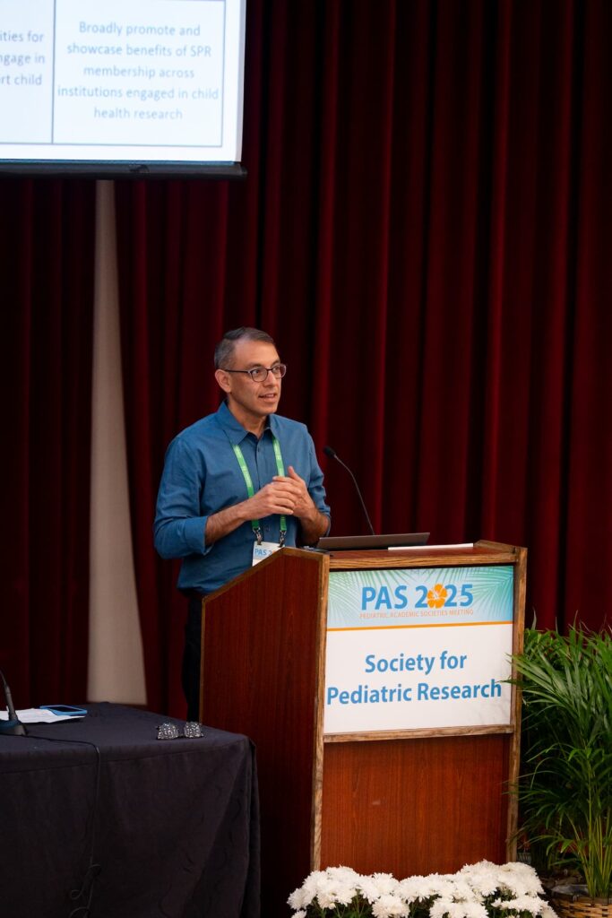 A person is standing at a podium labeled “PAS 2025 Society for Pediatric Research,” speaking in a conference setting. The background features dark red curtains, and there is a presentation screen above with partially visible text. White flowers and a green plant are placed near the podium.
