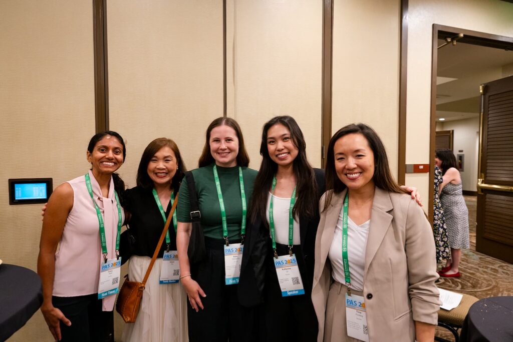 A speaker standing at a wooden podium labeled “Hawai‘i Convention Center,” delivering a presentation in a conference room. A banner to the left displays logos and text thanking sponsors, including the Children’s Hospital of Philadelphia. On the right, another person is seated at a table with several water bottles. Rows of chairs and attendees are visible in the foreground.