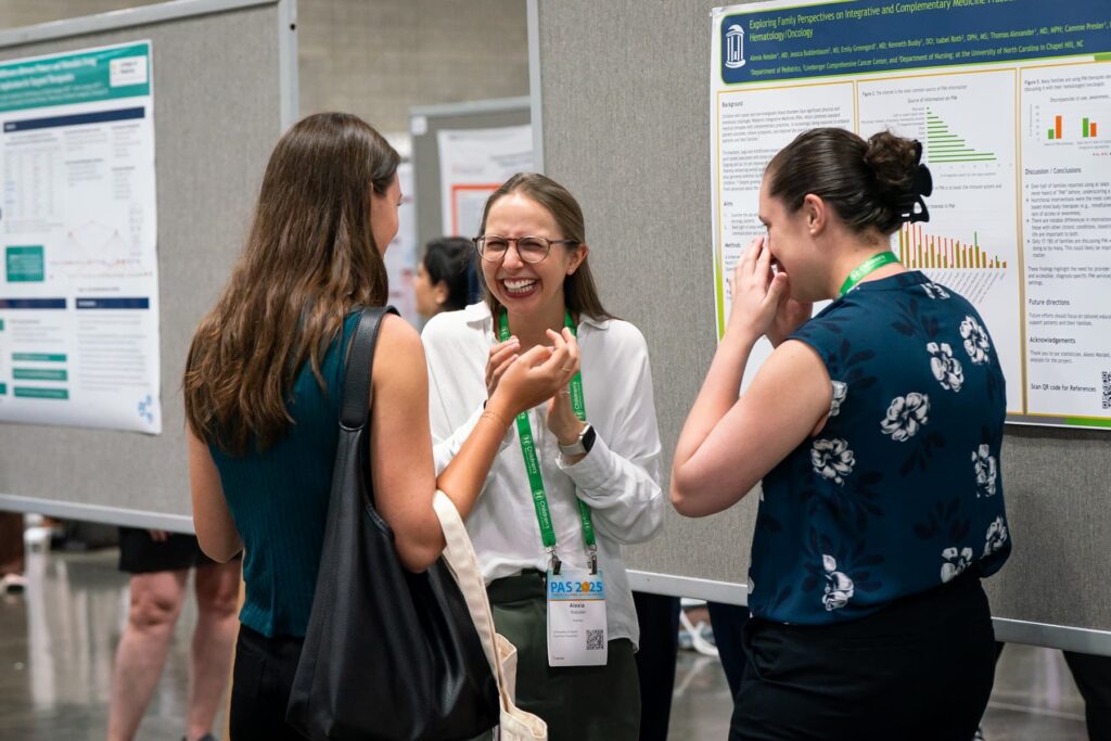 Three individuals standing and talking in front of research posters at a conference. The posters display charts, graphs, and text on gray boards. All participants are wearing event lanyards, and the setting appears to be an indoor exhibition area with polished flooring and multiple poster displays in the background.